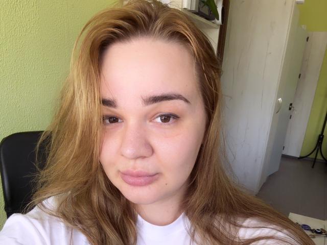 AmieAimee has light brown, slightly wavy hair, brown eyes, and a neutral expression with slightly puckered lips. She's wearing a white shirt, and her nose is slightly freckled.
