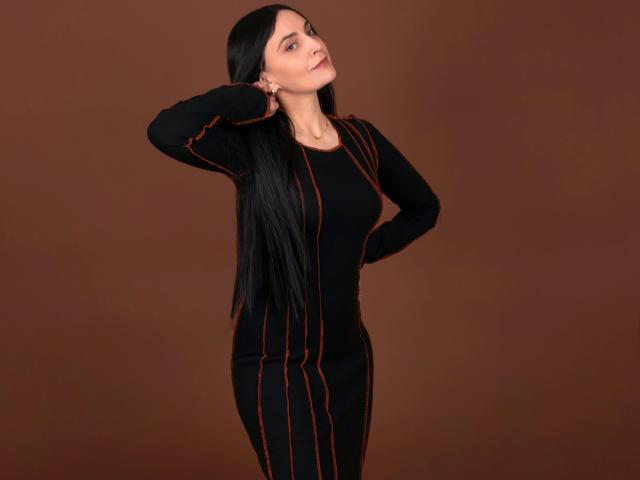 Xrystal-hot with long black hair, wearing a tight black dress with orange lines, hand on head, standing, brown background, looking slightly upward with a relaxed expression.