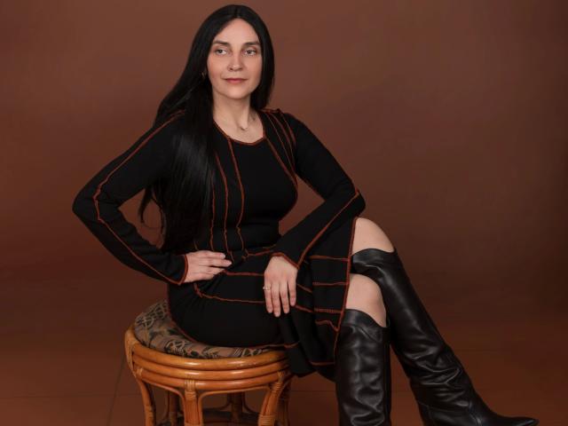 Xrystal-hot with long black hair, black dress with orange lines, and black boots, sitting on a wooden stool, smiling slightly, hands on her thigh, brown background.