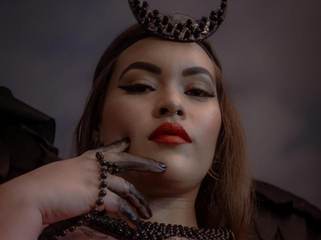 AlexaAblumm with dark eyebrows, red lipstick, and black beaded headpiece. She touches her face with black beaded fingers, wearing matching jewelry. Her red lips stand out.