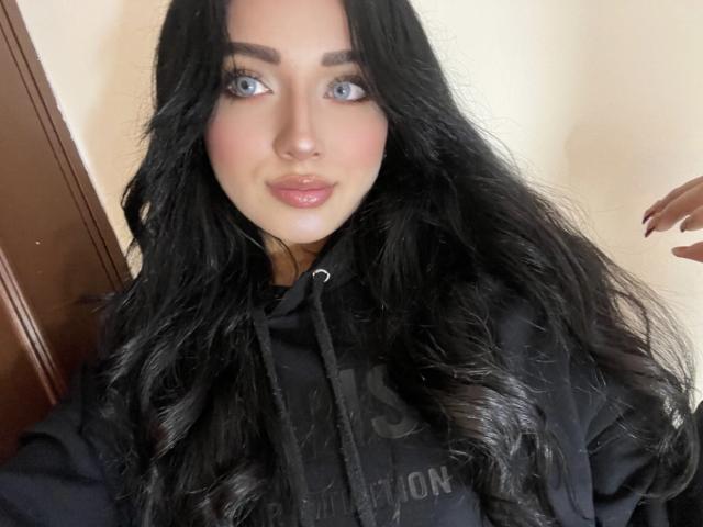 LillyTaylor with long black wavy hair, blue eyes, and glossy pink lips, wearing a black hoodie, showing a subtle smile, and holding a wooden door on the left.