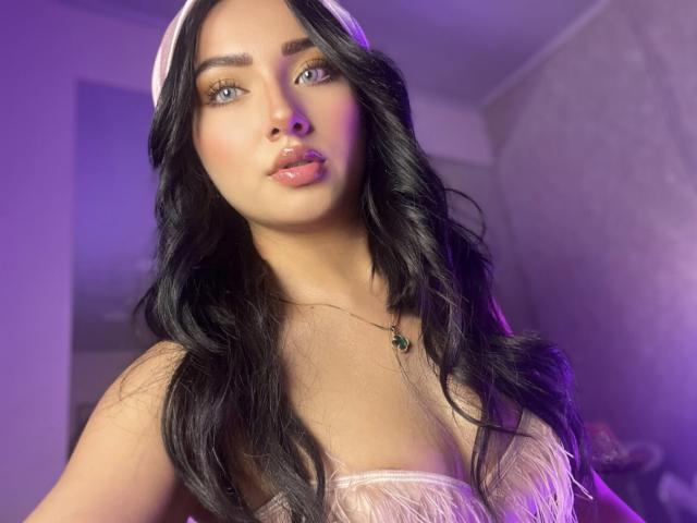 LillyTaylor with long black wavy hair, wearing a pink headband and sheer pink top, showing her tits, pink glossy lips, green necklace, purple lighting, and a cute expression.
