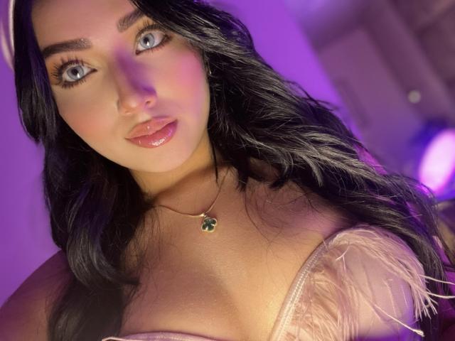LillyTaylor with long black hair, blue eyes, and pink lipstick, wearing a pink feathered top, showing her tits, and a green gem necklace, under purple lighting.