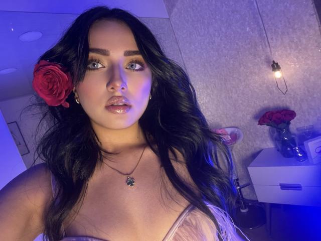 LillyTaylor with long black hair, red rose in her hair, blue eyes, glossy lips, wearing a pink dress, necklace with a green pendant, blue-lit room, hanging light bulb.