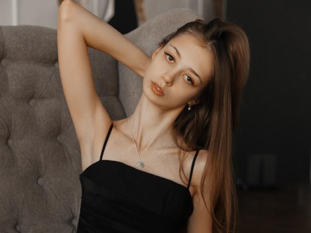 EmilyGraydis with long brown hair, wearing a black spaghetti strap dress, arms raised, looking seductively at the camera, showing her tits and a small silver necklace.