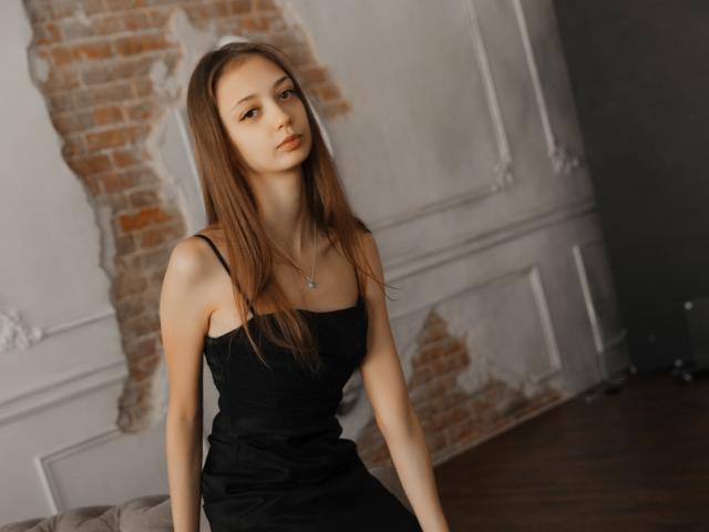 EmilyGraydis with long brown hair, wearing a black spaghetti-strap dress, sitting on a wooden floor, looking slightly to the side, with a subtle, confident expression.