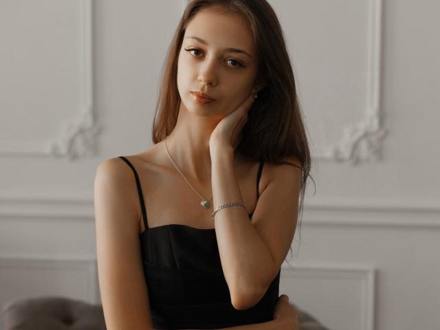 EmilyGraydis with long brown hair, wearing a black strap dress, touching her cheek, silver necklace, and bracelet, looking serious, soft light, elegant room, slim, small tits.
