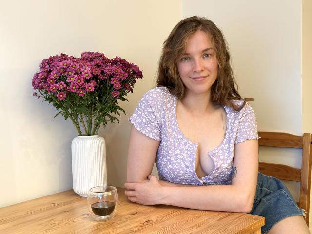 KenzieONEx with wavy brown hair, wearing a purple floral dress with a low neckline, showing a bit of her tits, sitting at a wooden table, glass of water.