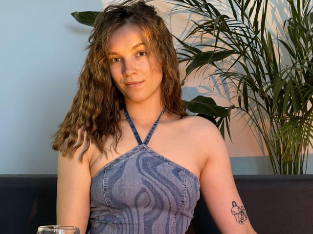 KenzieONEx has wavy brown hair, a small tattoo on her right arm, and is wearing a blue halter top with crisscross straps, showing her tits. She's smiling slightly.