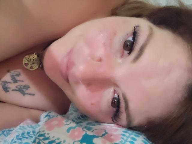 El rostro de Joyfull-hot está en primer plano, mostrando ojos marrones, una nariz rosada y una ligera sonrisa, con un pequeño tatuaje en su hombro derecho. Lleva un collar negro con un colgante dorado..