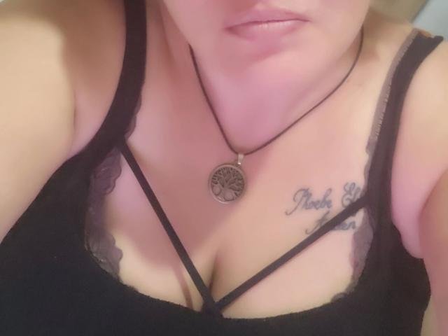 Het gezicht en de borst van Joyfull-hot, met een zwarte kanten BH, een ontwerp met bandjes, decolleté, een zwarte ketting met een ronde hanger, een tatoeage aan de rechterkant, lichtroze lippen, licht zichtbare armen, lichte huid..