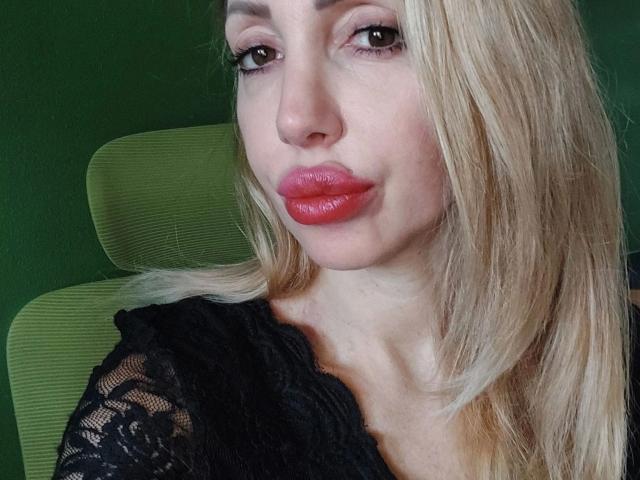LauraRubin-hot has blonde hair, red glossy lips, and black lace top, looking at camera with a slight smirk, green background visible.