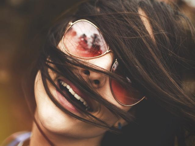 Saminaa's face with brown hair blowing across her pink-tinted sunglasses, smiling with visible teeth, her nose and mouth partially obscured, wearing a dark top, in a tight shot.