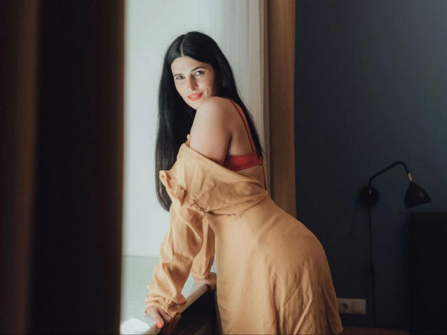 DarkWhiskers-hot with long black hair, red bra, and tan dress slipping off one shoulder, leaning forward, looking over shoulder, showing her ass, in a softly lit room.