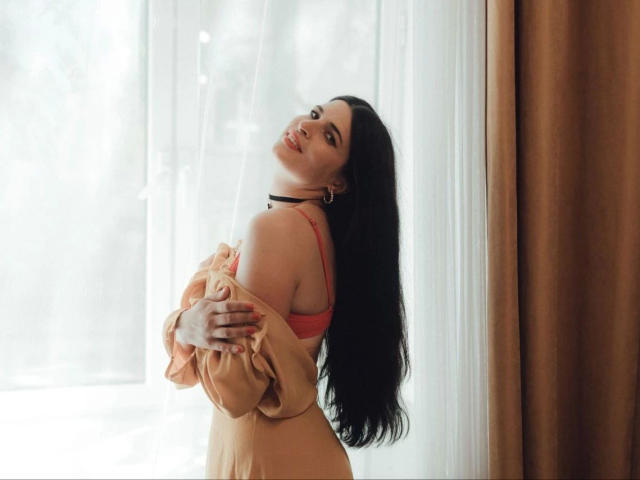DarkWhiskers-hot with long black hair, red bra, and beige off-shoulder top, smiling with head tilted, hands on chest, near window with brown curtain, looking sexy.