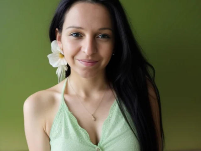 CatMelissa has long black hair, a white flower in her ear, and is wearing a green lace halter top, showing her small tits and a subtle smile.