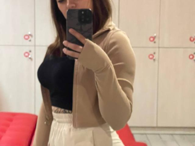 De selfie van Flimina toont haar in een beige jas, een zwarte top en een witte rok, terwijl ze een telefoon vasthoudt. Haar borsten zijn duidelijk zichtbaar in een kamer met rode meubels..