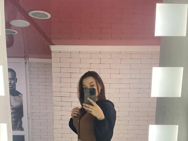 Flimina with brown hair, black top, and brown top, holding phone, mirror selfie, white brick wall, red ceiling, and muscular black-and-white poster in background.