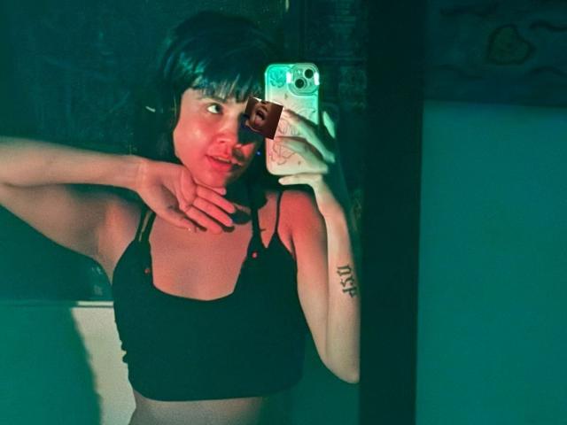 IsabellaMarynn with short black hair, wearing a black sports bra, holding a phone with a green case, taking a mirror selfie, showing a tattoo on her right arm.