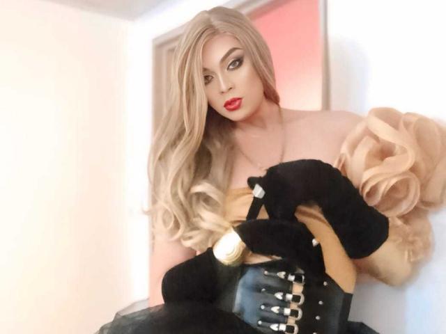 KarolElegance with long blonde hair, red lipstick, black gloves, tan puffy sleeves, black corset, holding black bag, looking seductively at camera, in a soft-lit room.