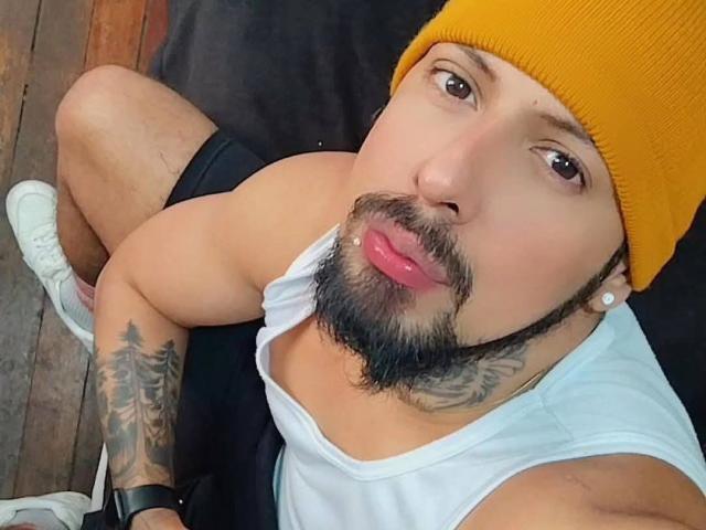JacobWalker, with a yellow beanie, white tank top, and black shorts, has a tattooed arm, beard, and is puckering up with glossy pink lips. Wearing white sneakers.