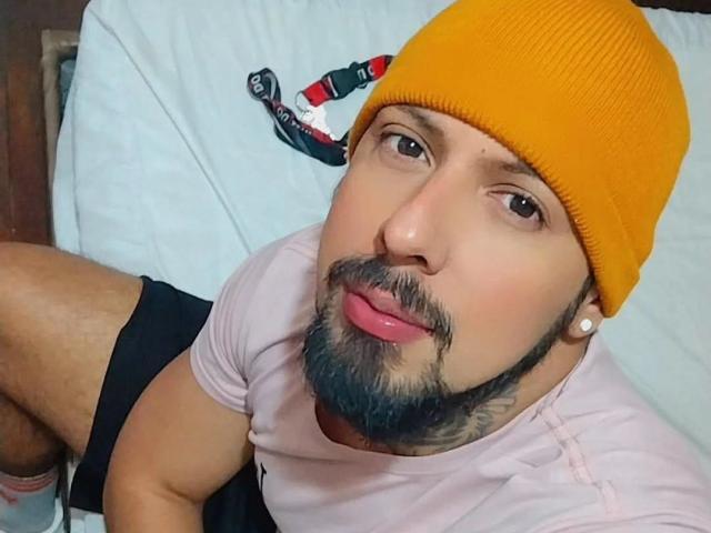 JacobWalker with a yellow beanie, pink t-shirt, black shorts, and a beard, sticking out his tongue, showing his pink ear studs, and looking up at the camera.