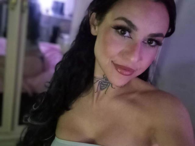 HappyBellaGio-hot with black hair, a tattoo under her neck, and a smile, wearing a strapless top, showing her bare shoulders and upper chest. Soft pink light in background.