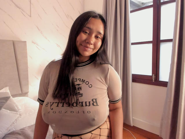Karollteyes with large, visible nipples through a sheer, tight, gray T-shirt, smiling at the camera, has long black hair, and is standing in a room with a window.