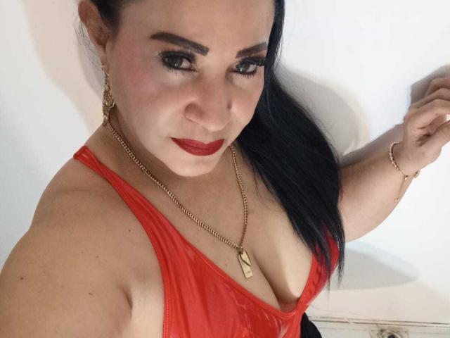 LucianaBlack, com batom vermelho, cabelo preto e um colar de ouro, vestindo um vestido vermelho brilhante que revela seus seios grandes. Ela está posando com um leve sorriso, olhando para a câmera..