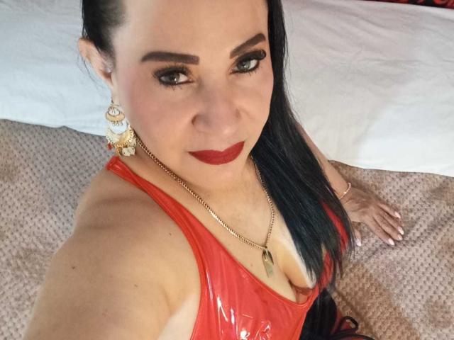 LucianaBlack trägt eine rote Latexbluse, hat glatte Haut, schwarzen Eyeliner, roten Lippenstift und langes schwarzes Haar. Sie trägt goldene Ohrringe und eine Halskette und blickt mit einem leichten Lächeln in die Kamera..