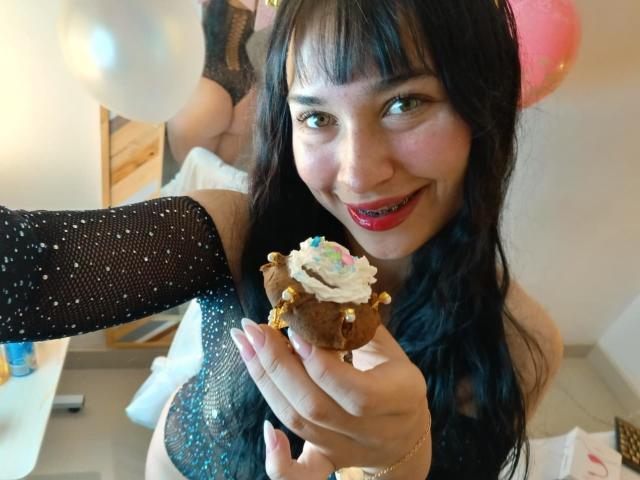 Sol Ortega, con lunghi capelli neri, rossetto rosso e una maglietta a rete, tiene in mano una cupcake con glassa bianca e codette colorate, sorridendo alla fotocamera e mostrando il suo viso carino..