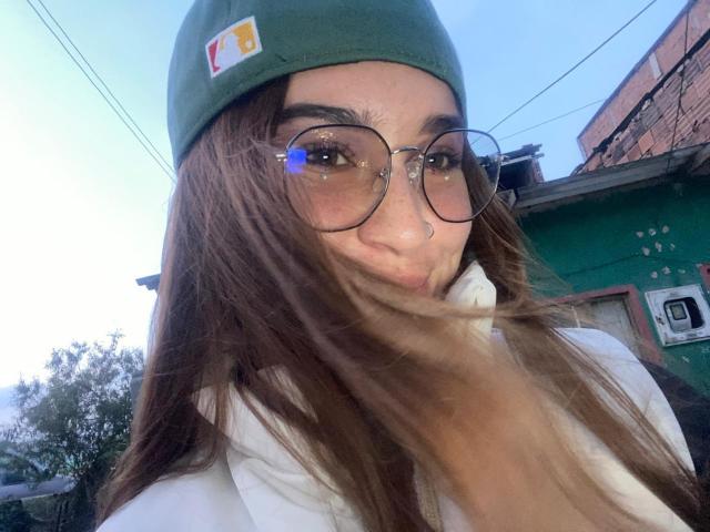 SolOrtega, avec de longs cheveux bruns, une casquette verte, des lunettes rondes, une chemise blanche, les lèvres pincées, un anneau au nez, et ses cheveux flottant sur le côté, sur une photo de rue..