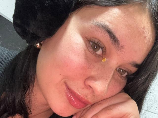 Le visage de SolOrtega, avec ses yeux marron foncé, sa peau légèrement rosée et un petit point doré au-dessus de son sourcil droit. Elle a des oreilles noires et duveteuses, des lèvres roses brillantes et un sourire détendu..