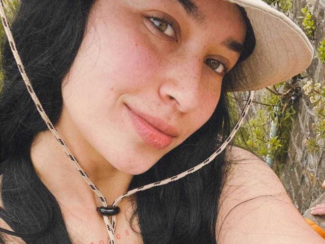 SolOrtega a une peau claire, des cheveux noirs ondulés et des yeux marron, avec un léger rougissement sur ses joues. Elle porte un chapeau beige et un collier à motifs, et elle sourit doucement..