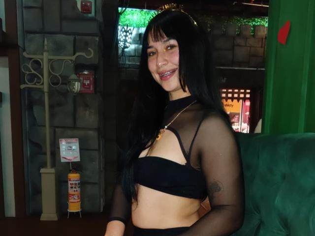 SolOrtega with long black hair, smiling, wearing a black sheer top that exposes her small tits and black bottom, sitting on a green couch.