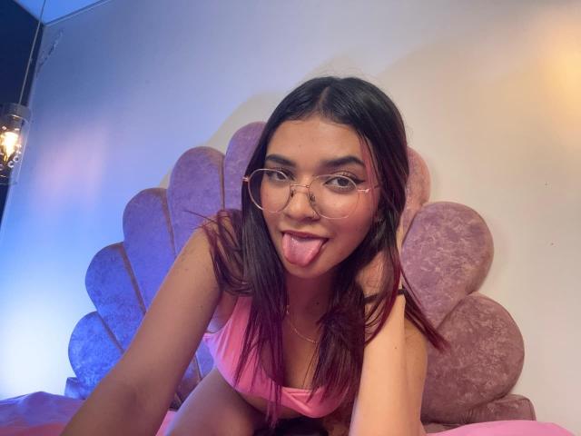 KiraRuby with glasses, tongue out, pink top, brown hair, and cute face, leaning on pink, plush, fan-shaped wall decoration. Her tits are visible. Bedroom setting.