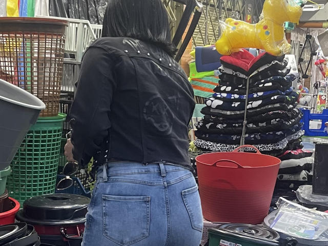 SamantaMuller with black jacket and blue jeans, standing with back to camera, showing her ass. Store has baskets, yellow stuffed dog, and folded clothes.