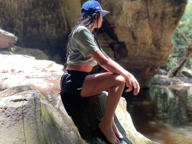 MiluSky, con trenzas largas, vistiendo una camiseta corta verde, pantalones cortos negros y una gorra azul, sentada sobre una roca, descalza, con las piernas cruzadas, en una zona rocosa y soleada..