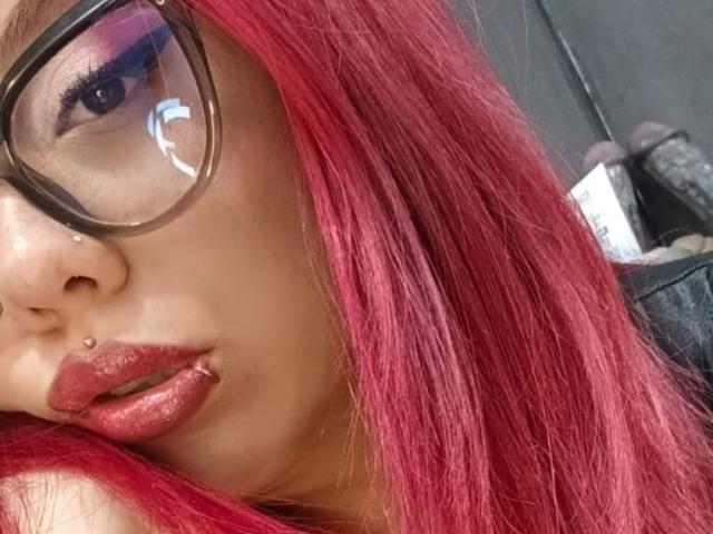 VelvetDomme con cabello rojo, gafas, piercings en la nariz, piercings en los labios y un lápiz labial rojo brillante, mirando ligeramente hacia un lado. Pecho parcialmente visible, bolso de cuero negro en el fondo..