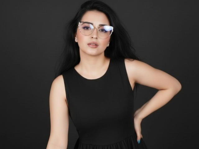 NicolJaen with long black hair, wearing a black sleeveless top, glasses, and blue nails, standing against a dark background, hands on hips, looking slightly to the side.