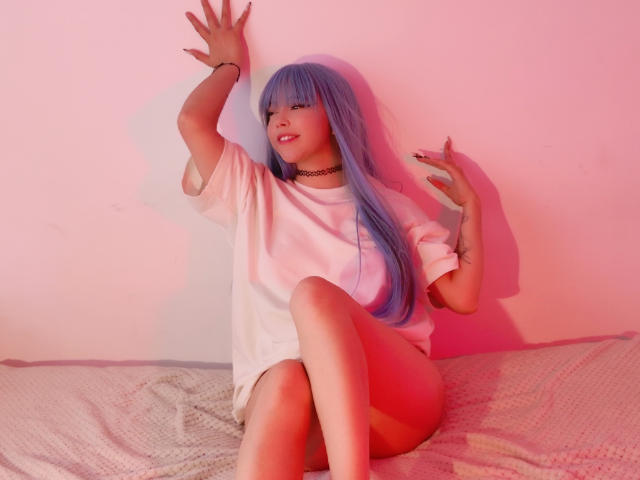 Arieyaara with blue hair, wearing a white tee and choker, sits on a bed with legs crossed, arms raised, and smiling. Pink and red light highlights her tits.