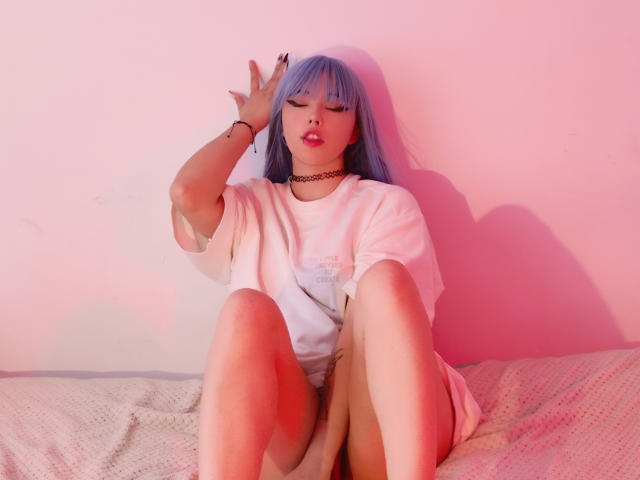 Arieyaara, com cabelo azul, vestindo uma camiseta branca e um colar preto, está sentada em uma cama com as pernas levantadas, a mão tocando sua cabeça, com iluminação rosa, mostrando seus seios..