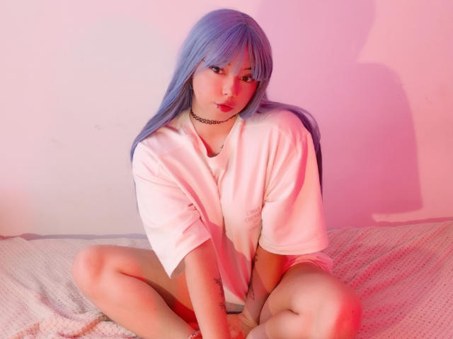 Arieyaara, avec des cheveux bleus, portant une chemise rose, assise les jambes écartées, montrant sa vulve, sur un lit avec un éclairage rose et rouge. Elle porte un collier ras du cou et regarde l'appareil photo..