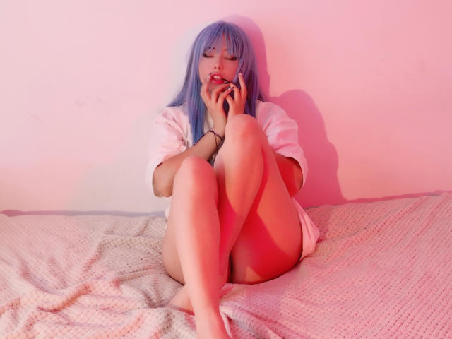 Arieyaara mit blauen Haaren, die Augen geschlossen, am Telefon, die Beine hochgelegt, trägt ein weißes Hemd und liegt auf einem pinkfarben beleuchteten Bett mit einer grauen, strukturierten Decke..