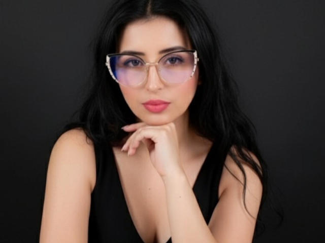 NicolJaen tiene el pelo largo y negro, usa gafas con un ligero tono morado y una camiseta negra sin mangas, con la mano cerca de sus labios rosados, mirando directamente a la cámara..