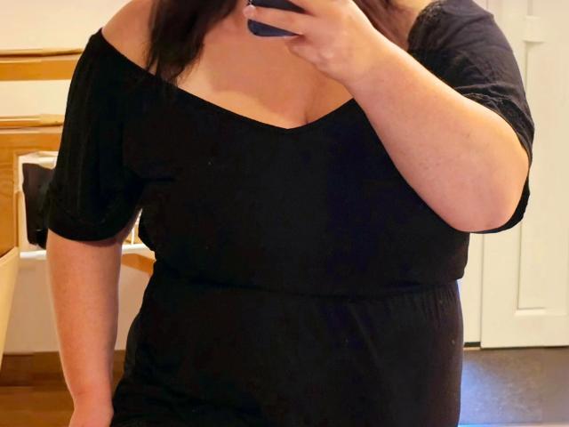 LADY-NORA-hot in a black off-shoulder dress, holding a phone to her face, showing her tits and a bit of her ass, with dark hair and fair skin.
