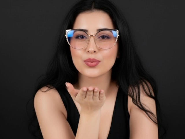 NicolJaen with long black hair, wearing clear glasses with blue reflections, pouting with pink lips, black sleeveless top, and hand near mouth, against a dark background.