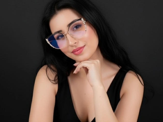 NicolJaen, con cabello negro, gafas de tono rosa y una blusa negra, mostrando su busto y una sonrisa encantadora, apoyando su barbilla en su mano..