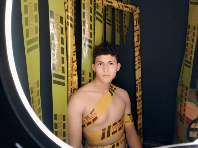 AxelSavage tiene el pelo negro y rizado y un tono de piel bronceado, vistiendo una cinta amarilla y negra a rayas que cubre su pecho y entrepierna, y está de pie frente a un aro de neón..