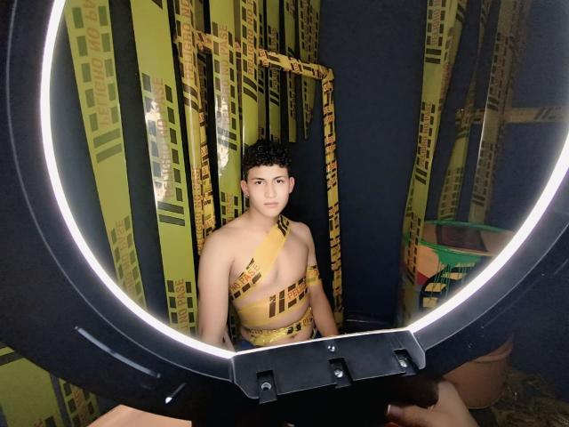 AxelSavage, met krullend zwart haar, draagt gele tape die hun borst en bovenlichaam bedekt, en zit voor een ronde ringlamp, waarbij ze recht in de camera kijken..